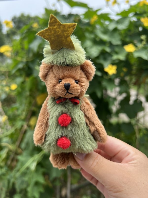 Knuffelbeer met kerstboomkleding en ster, vastgehouden in de hand met groen-bladerige achtergrond.