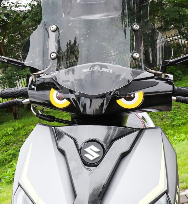 Zwarte Suzuki motor met opvallende gele oogstickers en windscherm, geparkeerd in groene omgeving.