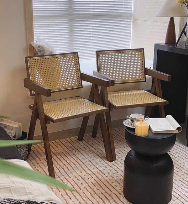 Twee houten rotan stoelen in een gezellige woonkamerhoek met klein tafeltje, boek en kaars voor een ontspannen sfeer.